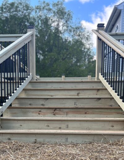 Wooden stairs with black metal railings leading to a deck. Trees and a partially visible house are in the background.